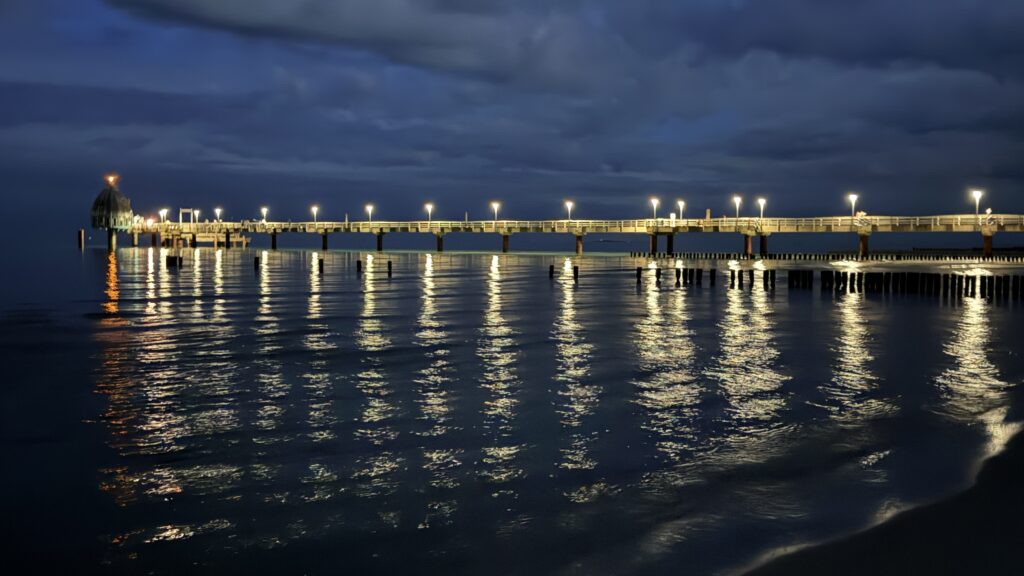 Ein langer Pier erstreckt sich über ruhiges, nächtliches Wasser, beleuchtet von Lichterketten, die sich auf der kräuselnden Oberfläche spiegeln. Die ruhige Szene fängt den Aufnahmeort eines Fotos ein, mit einer heiteren Atmosphäre, die durch den dunklen, bewölkten Himmel noch verstärkt wird.