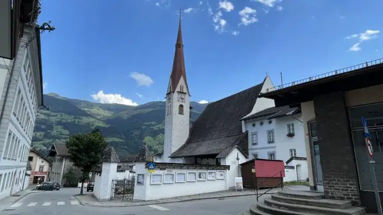 Ein kleines europäisches Dorf mit einer weißen Kirche mit hohem, spitzem Kirchturm und Uhr, umgeben von alpinen Bergen und traditionellen Gebäuden, ist der mögliche Aufnahmeort eines Fotos, das KI wie ChatGPT herausfinden könnte.