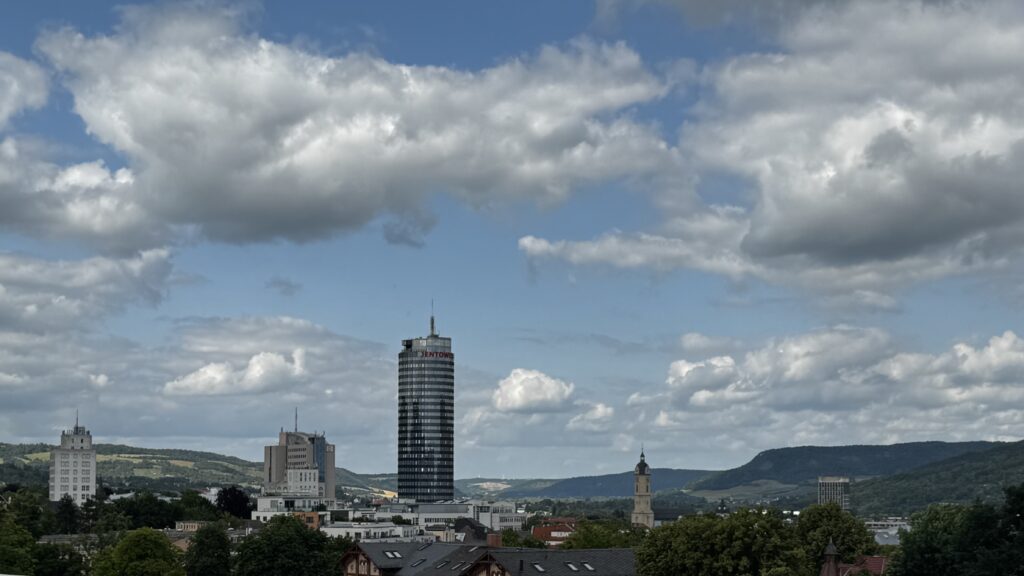 Stadtlandschaft mit einem hohen zylindrischen Wolkenkratzer in der Mitte, umgeben von kürzeren Gebäuden, grüne Bäume im Vordergrund - perfekt für einen ChatGPT Test zum Aufnahmeort eines Fotos. Im Hintergrund erheben sich Hügel unter einem teilweise bewölkten blauen Himmel.